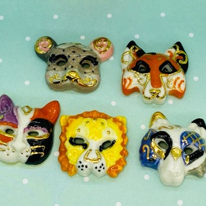 Op de afbeelding: Een verzameling van vijf keramische dierenmaskers, elk met unieke ontwerpen en kleuren. De maskers omvatten een luipaard, een tijger, een kat, een leeuw en een vos, allemaal met gouden accenten. Op een lichtblauwe achtergrond met witte stippen.