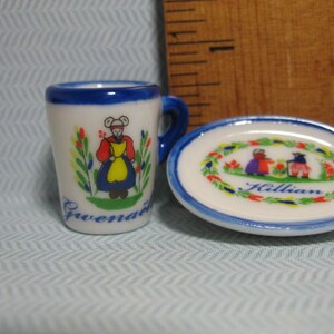 May include: A small white teacup with a blue rim and a painted design of a woman in a blue and yellow dress. The teacup has the word "Gwendal" written on the bottom. A small white saucer with a blue rim and a painted design of a wreath with red and yellow flowers and two people in blue and red dresses. The saucer has the word "Killian" written in the center.