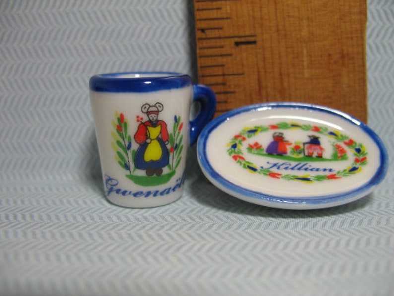 May include: A small white teacup with a blue rim and a painted design of a woman in a blue and yellow dress. The teacup has the word "Gwendal" written on the bottom. A small white saucer with a blue rim and a painted design of a wreath with red and yellow flowers and two people in blue and red dresses. The saucer has the word "Killian" written in the center.
