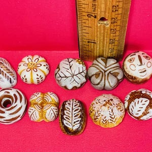 Puede incluir: Colección de diez esculturas de cerámica en miniatura de pan y pasteles. Las piezas están pintadas con detalles marrones y blancos, incluyendo un donut, una hogaza y una cara. Una regla de madera está en el fondo.