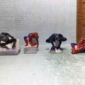 May include: Four small ceramic figurines of Spiderman in different poses. One is black and white, one is red and blue, and two are red and blue with black webbing.