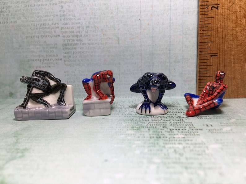 May include: Four small ceramic figurines of Spiderman in different poses. One is black and white, one is red and blue, and two are red and blue with black webbing.