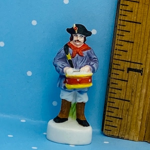 May include: A small porcelain figurine of a man wearing a blue jacket, red scarf, and black hat, playing a red and yellow drum. The figurine is standing on a white base.