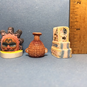 May include: Three small, colorful ceramic figurines. One features a sun-like face with a yellow base. The second is a brown vase with handles. The third is a rectangular structure with a detailed design. A wooden ruler is in the background.