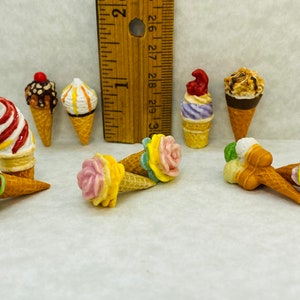 May include: A collection of miniature ice cream cones in various colours and flavours, including strawberry, chocolate, vanilla, and a floral design. The cones are made of ceramic and are approximately 5 cm tall.