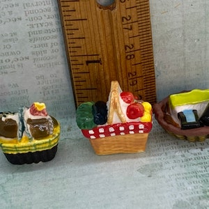 May include: Three miniature ceramic baskets filled with various items. The first basket is brown with a yellow and green plaid pattern and contains a brown loaf of bread, a yellow and white cake, and a small green and white item. The second basket is brown with a red and white checkered pattern and contains a green and black item, a red apple, and a yellow fruit. The third basket is brown and contains a green and yellow box, a blue and white box, and a brown bottle.