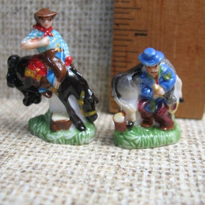 May include: Two miniature ceramic figurines depicting cowboys. One cowboy is riding a rearing black horse, while the other cowboy is standing next to a white horse. Both figurines are painted in bright colours and have detailed features.