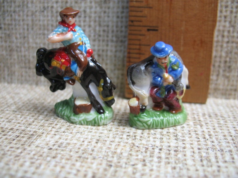 May include: Two miniature ceramic figurines depicting cowboys. One cowboy is riding a rearing black horse, while the other cowboy is standing next to a white horse. Both figurines are painted in bright colours and have detailed features.