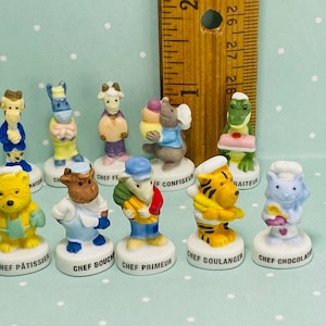 May include: A set of ten miniature figurines of animals dressed as chefs. Each figurine is holding a different food item, such as a cake, a loaf of bread, or a pastry. The figurines are all painted in bright colors and have a whimsical, cartoonish style. The figurines are labeled with the names of different types of chefs, such as "Chef Pâtissier", "Chef Boulanger", and "Chef Confiseur".