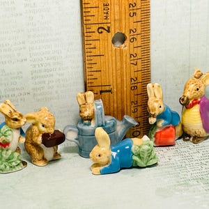 May include: A collection of five miniature ceramic figurines of rabbits and a watering can. The rabbits are wearing blue and purple clothing and holding various objects, including a watering can, a book, and a basket. The figurines are painted in a realistic style and have a vintage look.