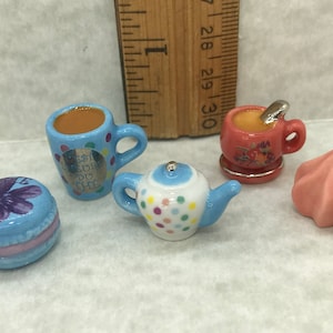 Puede incluir: Un juego de tazas de té y tetera en miniatura, que incluye una tetera de lunares azules y blancos, una taza de té azul y blanca con el texto "Best Friends Do Tea", una taza de té rosa con diseños florales, un macaron rosa con un diseño de flor morada y una galleta de merengue rosa.
