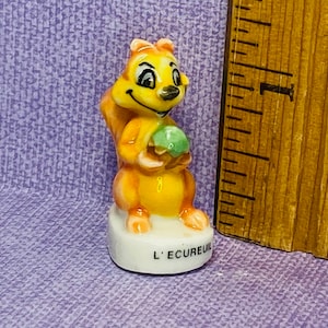 May include: A small ceramic figurine of a squirrel holding a green acorn. The figurine is painted in shades of yellow and orange. The base of the figurine is white and has the words "L'Ecurie" written on it.