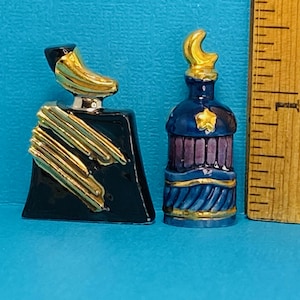 Puede incluir: Dos pequeñas botellas decorativas con detalles dorados. La botella de la izquierda es negra con rayas doradas y una parte superior en forma de media luna. La botella de la derecha es azul con detalles morados y dorados y una parte superior en forma de media luna.