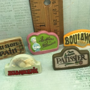 Könnte beinhalten: Fünf Miniatur-Keramikschilder mit französischen Bäckereimotiven. Die Schilder sind in verschiedenen Farben gehalten und zeigen französischen Text, darunter "Au Bon Pain", "Boulangerie", "Les Delices du Patissier", "Patissier" und "Boulanger".