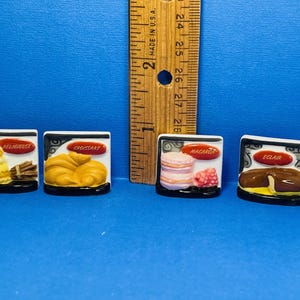 Könnte beinhalten: Vier kleine Keramikfliesen mit Bildern verschiedener französischer Gebäcke. Die Fliesen sind weiß mit schwarzen Rändern und rotem Text. Die Gebäcke sind ein Mille-feuille, ein Croissant, Macarons und ein Éclair.