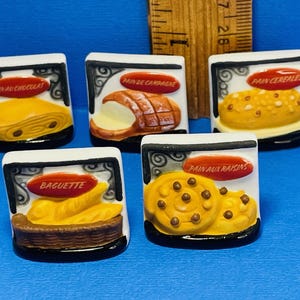 Könnte beinhalten: Fünf Keramikfliesen mit Bildern von verschiedenen Brotsorten. Die Fliesen sind weiß mit schwarzem Rand und zeigen roten Text mit den Namen der Brote: "Pain au Chocolat", "Pain de Campagne", "Pain Cereales", "Baguette" und "Pain aux Raisins".