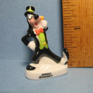 May include: A small porcelain figurine of Goofy dressed in a black tuxedo with a green vest and a top hat. He is holding a cane in his right hand and has a bow tie around his neck. The figurine is on a white base with the words "DINGO ©DISNEY" printed on it.