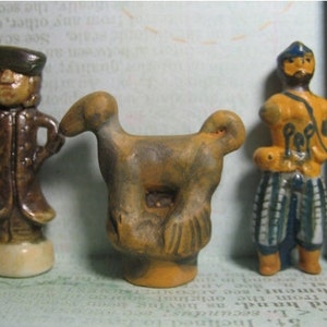 May include: Three small ceramic figurines: a man in a brown coat and hat, a brown and yellow bird, and a man in a yellow and blue outfit. The figurines are painted with details and have a rustic look.