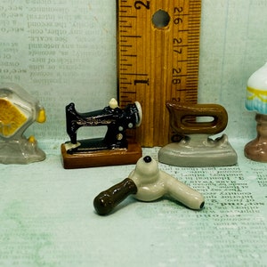 Op de afbeelding: Vijf miniatuur keramische beeldjes die huishoudelijke artikelen voorstellen: een naaimachine, een strijkijzer, een föhn, een lamp en een vintage radio. De beeldjes zijn in verschillende kleuren geschilderd, waaronder zwart, wit, bruin, blauw en geel.