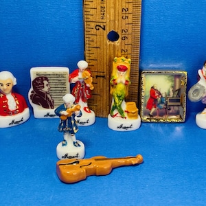 May include: A collection of miniature porcelain figurines depicting Wolfgang Amadeus Mozart. The figurines include a bust, figures playing instruments, and a framed painting, all with the name "Mozart" on the base. A wooden ruler is in the background.