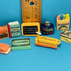 May include: A collection of miniature school supplies, including a brown briefcase, a blue ink bottle, a yellow pencil box, a green book with the text "LANGUE FRANCAISE ABCD", a green book with the text "BON-POINT", a yellow book with the text "Plumier", a black chalkboard with the text "J'écouteraix la maîtresse en classe.", and a yellow and white box with the text "COULEUR".
