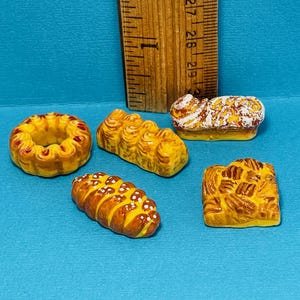 May include: Five miniature pastries, including a ring-shaped pastry, a loaf of bread, a braided loaf, a square pastry, and a long loaf. The pastries are all a light brown colour and have a glazed finish.
