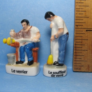 May include: Two ceramic figurines depicting a glass blower and a glass maker. The glass blower is wearing a white shirt and blue pants and is holding a glass blowing tool. The glass maker is wearing a brown shirt and blue pants and is sitting at a workbench. The figurines are on a white base with the text "Le souffleur de verre" and "Le verrier" written on them.