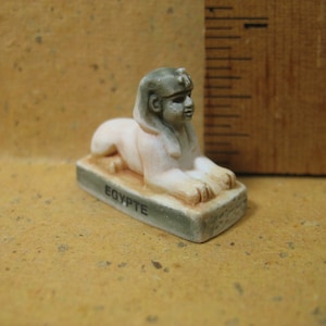 May include: A miniature sphinx figurine with a grey head and white body, resting on a rectangular base labeled EGYPTE. The figurine is positioned next to a wooden ruler, showing the object is approximately 0.75 inches in height at the head.