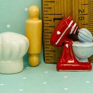 Może przedstawiać: Zestaw trzech miniaturowych przedmiotów kuchennych z ceramiki: biały czepek kucharza, drewniany wałek do ciasta i czerwono-biała miksera stojąca z niebieską misą.