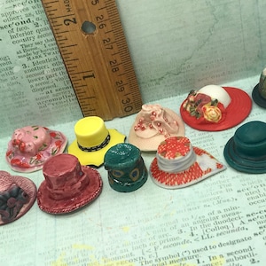 May include: A collection of ten miniature ceramic hats in various colors and patterns, including pink, red, yellow, green, and blue. The hats are arranged on a surface with a ruler for scale.