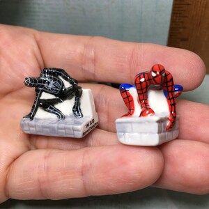 May include: Two small ceramic figurines of Spiderman characters. The figurine on the left is black and the figurine on the right is red and blue. Both figurines are standing on small white platforms. The text "TM & &copy; Marvel Characters" is printed on the platform of the black figurine.