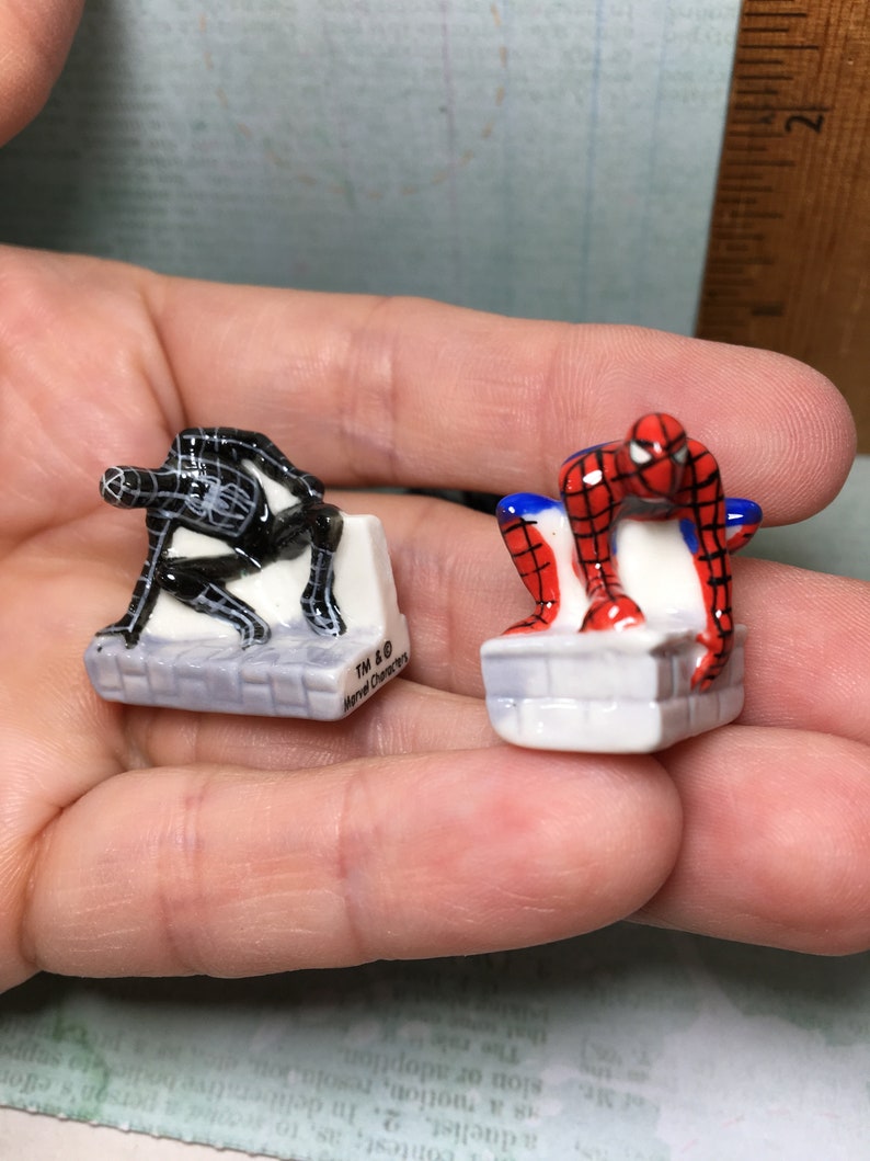 May include: Two small ceramic figurines of Spiderman characters. The figurine on the left is black and the figurine on the right is red and blue. Both figurines are standing on small white platforms. The text "TM & &copy; Marvel Characters" is printed on the platform of the black figurine.