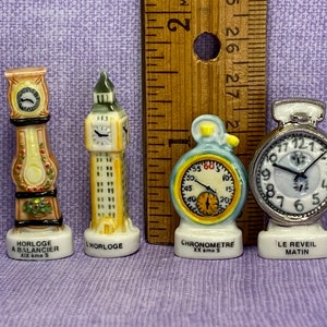 May include: Four miniature porcelain clocks, each with a different design. The first is a tall, ornate clock with a floral pattern. The second is a tower clock with a white face and black hands. The third is a round clock with a yellow face and black hands. The fourth is a round clock with a white face and black hands. The clocks are labelled "HORLOGE A BALANCIER XIX ème S", "LHORLOGE", "CHRONOMETRE XX ame S", and "LE REVEIL MATIN".