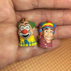 May include: Two ceramic clown figurines. One clown has a green and yellow hat, a red nose, and a blue and white checkered scarf. The other clown has a multi-colored jester hat and a purple and gold collar.