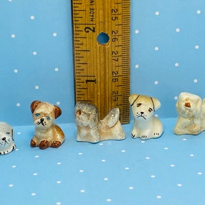 Może przedstawiać: Pięć małych ceramicznych figurek psów, każda w innej pozie i z malowanymi detalami. Figurki są białe, brązowe i beżowe. Linijka znajduje się w tle jako odniesienie do rozmiaru.