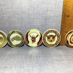 May include: A collection of five small, round decorative plates. Each plate features a different African animal: a crocodile, elephant, antelope, leopard, and lion. The plates have a cream-colored background with brown and gold accents.