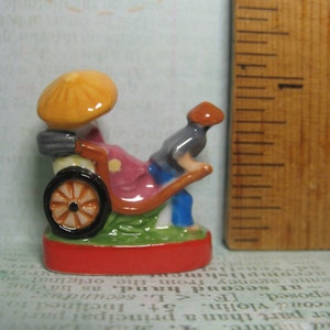 May include: A miniature ceramic figurine of a person riding a rickshaw. The rickshaw is yellow, red, and green with a black wheel. The person is wearing a blue shirt and a red hat.
