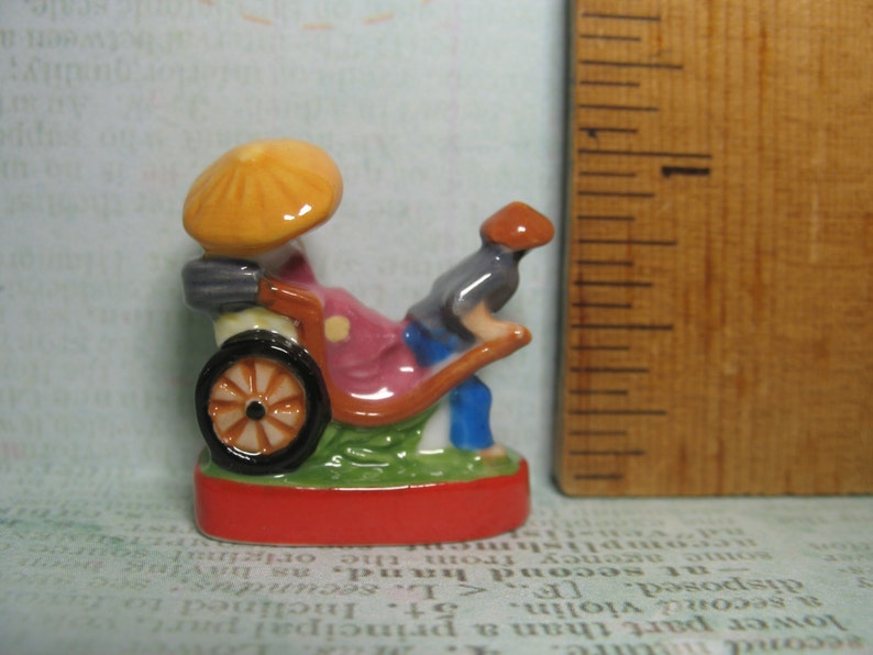 May include: A miniature ceramic figurine of a person riding a rickshaw. The rickshaw is yellow, red, and green with a black wheel. The person is wearing a blue shirt and a red hat.