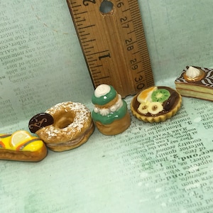Peut inclure: Six miniatures de pâtisseries en céramique, dont un beignet saupoudré de sucre glace, une pâtisserie verte et blanche avec un motif en spirale, une tarte aux fruits et au chocolat, une pâtisserie au citron, une pâtisserie avec un motif en spirale chocolat et blanc, et une pâtisserie avec un motif en spirale brun et blanc.