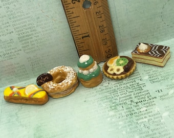 May include: A collection of six miniature ceramic pastries, including a donut with powdered sugar, a green and white pastry with a swirl design, a tart with fruit and chocolate, a pastry with a lemon design, a pastry with a chocolate and white swirl design, and a pastry with a brown and white swirl design.