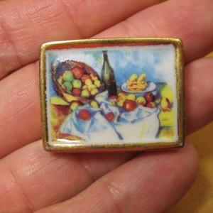 Puede incluir: Un broche rectangular con marco de metal dorado que presenta una escena de bodegón impresa a color con un cuenco de frutas, una botella oscura y un paño blanco sobre una mesa. La obra muestra tonos vibrantes de rojo, amarillo y verde.