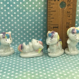May include: Four small ceramic unicorn figurines. The unicorns are white with rainbow colored manes and tails. One unicorn is lying down, one is standing with its head down, and two are standing upright.