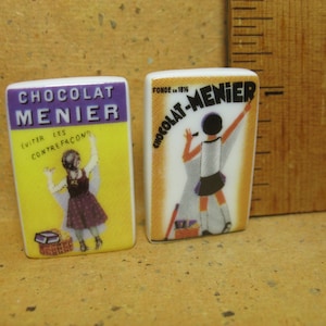 Puede incluir: Dos azulejos rectangulares de cerámica con anuncios antiguos de Chocolat Menier. El azulejo izquierdo es amarillo con texto morado y una niña con vestido. El derecho es crema con texto negro y una niña pintando. Una regla muestra 2,5 cm.