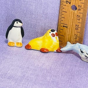 Könnte beinhalten: Eine Sammlung von Miniatur-Keramikfiguren, darunter ein Pinguin, eine Robbe und ein Wal. Der Pinguin ist schwarz-weiß, die Robbe gelb mit roten Akzenten und der Wal hellblau. Ein Holzlineal ist im Hintergrund.