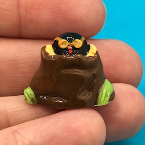 May include: A small, hand-painted figurine of a black creature with yellow goggles and a red beak, emerging from a brown sack. The figurine has green accents at the base. The overall design is whimsical and detailed.