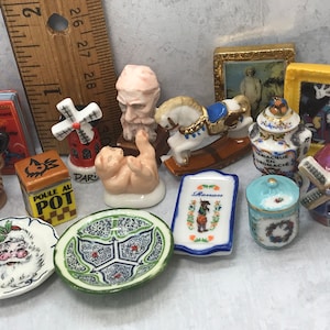 May include: A collection of miniature ceramic figurines, including a windmill, a baby, a rocking horse, and a small painting. Other items include a small book, a plate with a Santa Claus design, and a small bowl with a floral pattern.