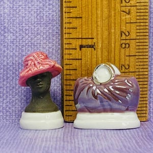 May include: Two miniature ceramic figurines. The first figurine is a woman wearing a pink hat and a black dress. The second figurine is a woman wearing a purple dress and a white hat.