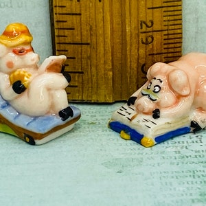 May include: Two ceramic pig figurines. One pig is wearing a hat and sunglasses and is sitting in a blue and white lounge chair. The other pig is wearing a mustache and is lying on a blue and white book.