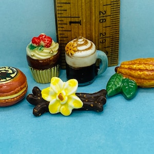 May include: A collection of miniature ceramic figurines depicting various desserts and spices. The set includes a chocolate cupcake with red and green berries, a cup of coffee with whipped cream, a vanilla bean pod with a yellow flower, a cocoa pod with green leaves, and a red and brown macaron with a gold design.