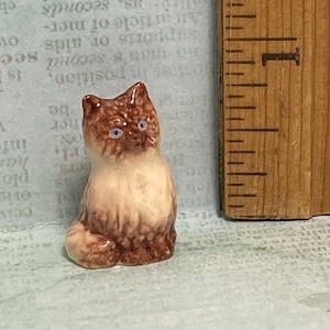 以下が含まれることがあります： 青い目をした茶色と白の猫の小さなセラミック製フィギュア。猫は尻尾を脚に巻き付けて座っています。